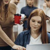 A woman is sitting at her laptop while another person stands nearby.