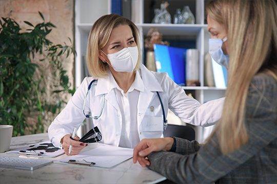 A woman in white lab coat and mask talking to another person.